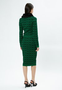 Grünes gemustertes Kleid mit langenÄrmeln, ausgestattet mit einem schwarzen Pelzkragen. Figurnah geschnitten, knielanges Design mit Zickzackmuster. Stilettos.