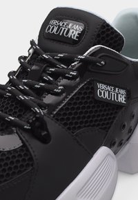 Μαύρο αθλητικό παπούτσι Versace Jeans Couture με κορδόνια με σχέδιο, πλευρές από δίχτυ μέλισσας και λεπτομέρειες λευκής λαστιχένιας σόλας.