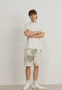 White cotton t-shirt with a crew neckline, paired with light beige shorts featuring a grid pattern. White sneakers and beige socks complete the look.