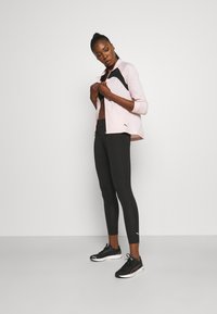 Helle rosa Zip-Jacke mit schwarzen Akzenten, getragen über einem schwarzen Top, kombiniert mit schwarzen Leggings und schwarzen Sneakers. Glatter Stoff, athletischer Schnitt.