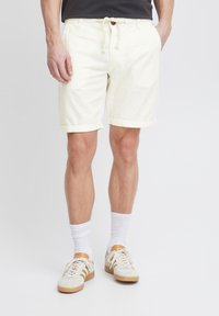 Pantaloni scurți din amestec de bumbac, de culoare galben deschis, cu talie ajustabilă prin șnur și manșete răsucite, asortați cu șosete albe și adidași bej.