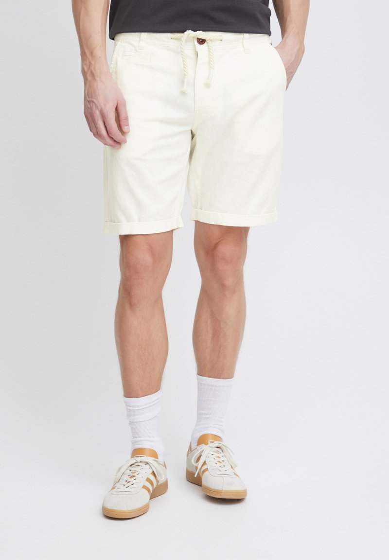 Pantaloni scurți din amestec de bumbac, de culoare galben deschis, cu talie ajustabilă prin șnur și manșete răsucite, asortați cu șosete albe și adidași bej.