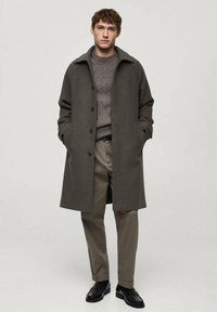 Gray long coat with a collar, button front, and side pockets, layered over a textured gray sweater and beige trousers, worn with black shoes.