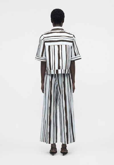 Άτομο που φαίνεται από πίσω φορά κοντομάνικο σακάκι και φαρδιά παντελόνια με κάθετες μαύρες και λευκές ρίγες, στέκεται σε λευκό φόντο.