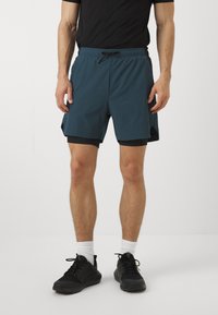 Shorts deportivos en color teal con un forro interior negro, cintura elástica y bolsillos laterales. Llevado con zapatillas negras y calcetines blancos.