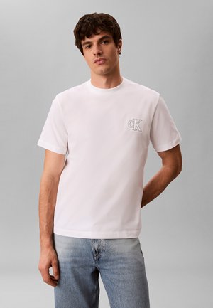 Giovane uomo con t-shirt bianca a maniche corte con piccolo logo "cK" sul petto e jeans azzurri chiari, in piedi davanti a uno sfondo grigio.