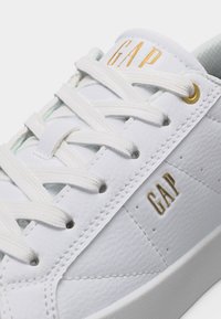 Baskets en cuir blanc avec des lacets blancs et des logos "GAP" dorés sur la languette et sur le côté près des œillets, vue en gros plan.