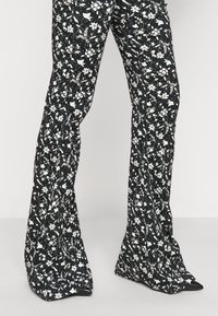 Svarta utsvängda byxor med ett vitt blommönster, tillverkade av ett lätt tyg med en slät yta och spetsiga svarta skor.