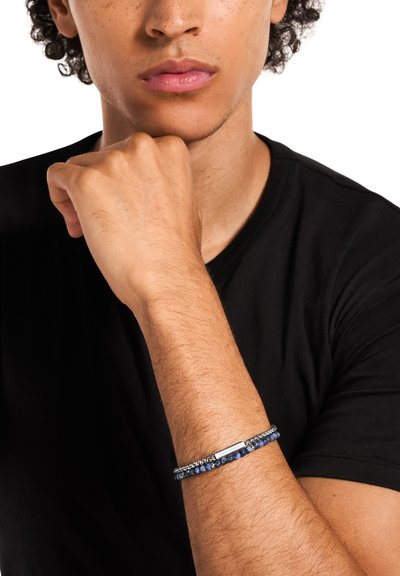 Bracelet bleu marine orné de perles avec des éléments en argent, présentant des pierres lisses et texturées. Porté au poignet, associé à un t-shirt noir.