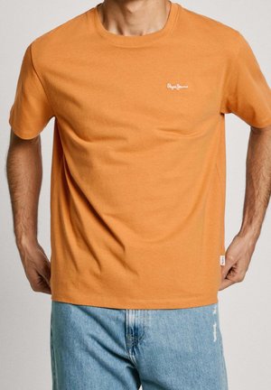 Man in oranje T-shirt met korte mouwen en klein wit "Pepe Jeans" logo op de borst, en lichtblauwe, versleten jeans. Handen in de zij.