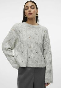 Maglione grigio chiaro a maglia con motivo a treccia e dettagli a taglio, scollo rotondo e maniche leggermente accorciate. Indossato sopra pantaloni grigi.