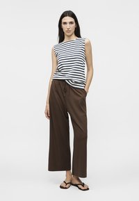 Femme portant un haut sans manches à rayures bleu marine et blanches, un pantalon ample marron et des tongs noires, debout devant un fond clair uni.