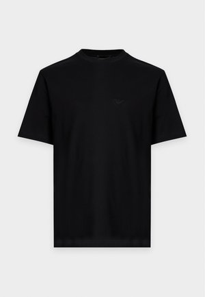Junger Mann steht, trägt ein schwarzes Kurzarm-T-Shirt, beige weite Hosen und schwarze Schuhe vor einem schlichten hellen Hintergrund.