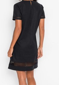 Schwarzes Kleid mit kurzen Ärmeln, das einen strukturierten gewebten Saum am Rand und an den Ärmeln sowie eine Schlüsselöffnung am Rücken aufweist.