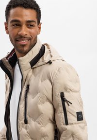 Doudoune beige avec capuche, dotée de poches zippées, d'accents noirs et d'un matériau texturé. L'intérieur présente des couleurs contrastantes.