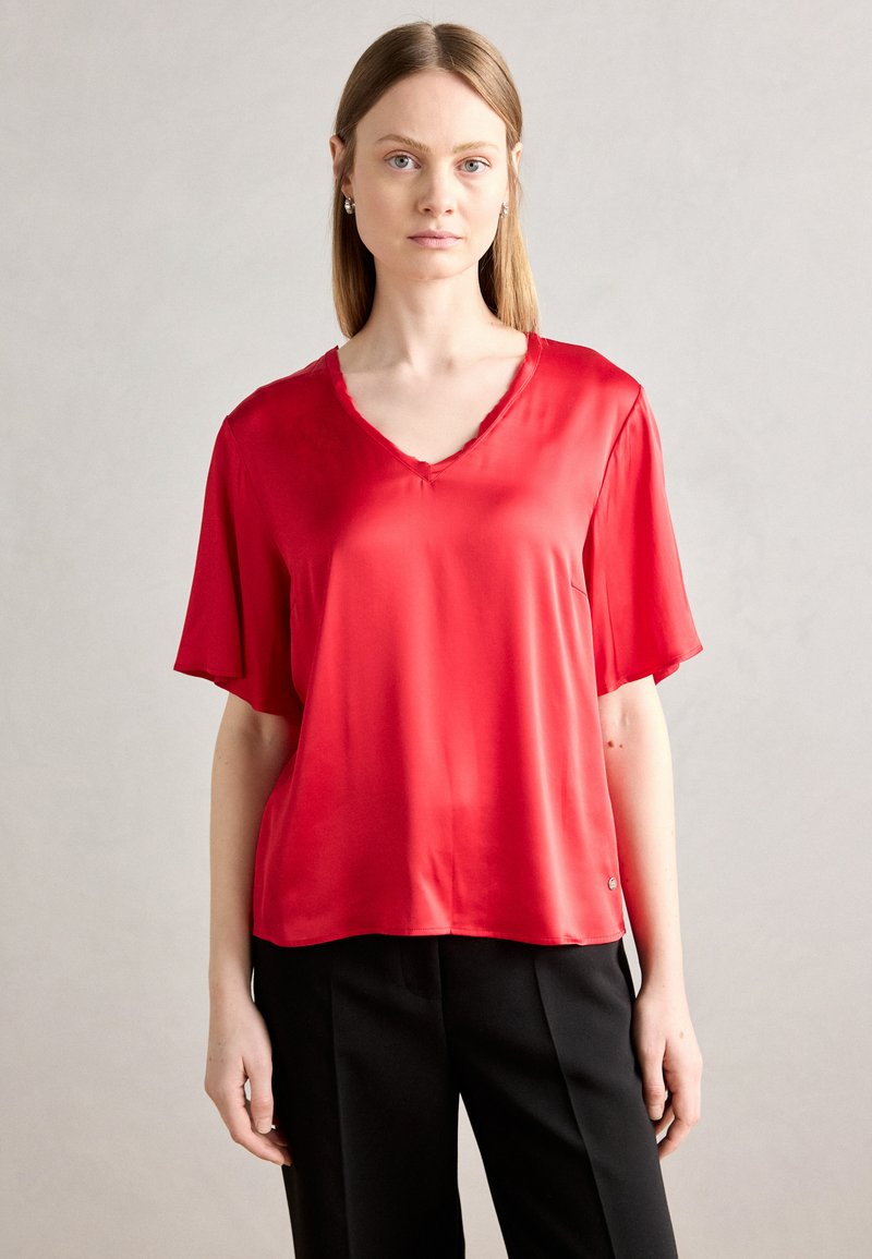 Blusa corta in satin rosso a maniche corte con scollatura a V e vestibilità rilassata, caratterizzata da un dettaglio logo laterale e una superficie liscia su uno sfondo neutro.