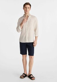 Jasnobeżowa lniana koszula, granatowe szorty i czarne sandały. Koszula ma zapięcie na guziki przy kołnierzyku i podwinięte rękawy, charakteryzuje się luźnym krojem.