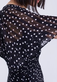 Czarna sukienka w białe groszki z przezroczystymi, falbanowymi rękawami, elastyczną talią i kontrastującymi białymi kropkami. Lekka, zwiewna tkanina o teksturowanym wyglądzie.