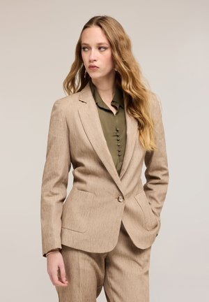 Ung kvinde med langt, bølget hår iført en beige dragt og en olivengrøn knapbluse, der kigger mod venstre med hænderne i lommerne.