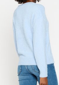 Pull bleu clair duveteux avec un col arrondi, des manches longues et un ourlet côtelé. Porté avec un jean bleu ajusté. Texture douce visible.