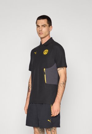 Sort polo skjorte med gule detaljer, korte ærmer og en lynlåslomme. Har et gult cirkulært logo på brystet og et mærke emblem.
