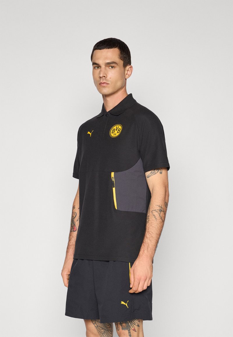 Sort polo skjorte med gule detaljer, korte ærmer og en lynlåslomme. Har et gult cirkulært logo på brystet og et mærke emblem.