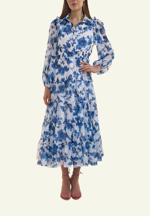 Frau trägt ein weißes Maxi-Kleid mit langen Ärmeln und blauem Blumenmuster, gestufter Rock und pinke, geschnürte High Heels, steht mit einer Hand in der Hüfte.