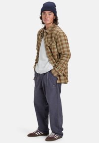 Braunes kariertes Flanellhemd über einem grauen Rundhalsausschnitt, kombiniert mit marineblauen Cargohosen und braunen Sneakers mit weißen Streifen. Mütze auf dem Kopf getragen.