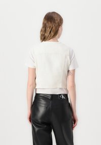 Kuttet kremfarget denimvest med en tettsittende design, vist over en hvit t-skjorte. Kombinert med svarte skinnbukser med høy midje som har et logobelte.