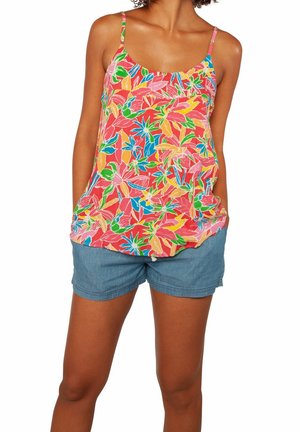 Femme portant un débardeur coloré à fleurs avec des bretelles fines et un short en jean bleu, debout les mains dans les poches.