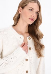Kötött fehér kardigán texturált mintákkal, kerek nyakkivágással és barna gombokkal. Puha, bolyhos anyag, részletes díszítéssel az ujjakon.