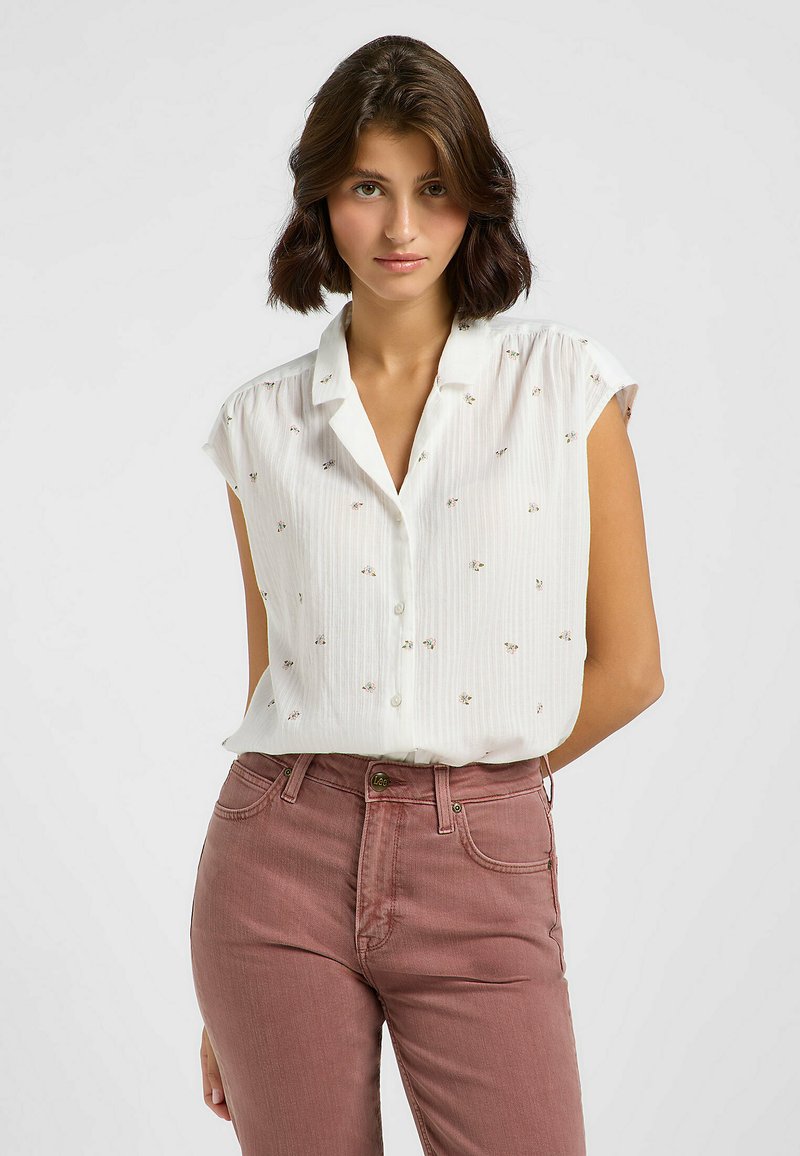 Camisa blanca de manga corta con rayas verticales y bordados florales dispersos, combinada con unos jeans color rosa empolvado con cierre de botón.