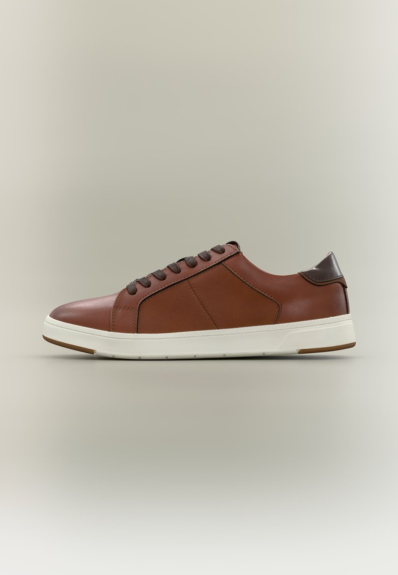 Καφέ δερμάτινο αθλητικό παπούτσι χαμηλού προφίλ με σκουρόχρωνα καφέ κορδόνια, υφασμένο πλαϊνό πάνελ, λευκή σόλα και σκουρόχρωμη καφέ ταμπέλα στη φτέρνα σε ουδέτερο φόντο.