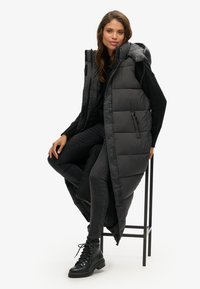 Schwarze Steppweste mit langem Design, Reißverschluss und Kapuze. Verfügt über große gestepfte Abschnitte und eine Seitentasche. Getragen über einem schwarzen Oberteil und Jeans.
