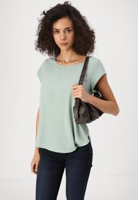 Jauna sieviete ar čirkainiem matiem, ģērbusies gaiši zaļā īsām piedurknēm topā un tumšās džinsās, tur brūnu plecu somu uz vienkrāsaina fona.