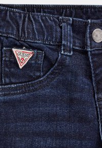 Jeans de mezclilla azul oscuro con un acabado texturizado, que presentan un parche triangular rojo y blanco con el logo "GUESS" en el bolsillo frontal y un botón metálico.