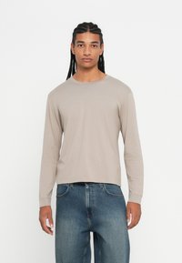 Jonge man met gevlochten haar draagt een beige shirt met lange mouwen en een blauwe spijkerbroek, staand tegen een effen witte achtergrond.