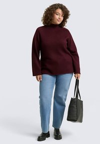 Bordowy sweter z wysokim kołnierzem i długimi rękawami, zestawiony z jasnoniebieskimi dżinsami i czarnymi botkami na platformie, trzymający czarną torbę typu tote.