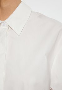 Bílá košile s límečkem vyrobená z hladké, lehké látky s čistým vzhledem; obsahuje zapínání na knoflíky a decentní texturové detaily.