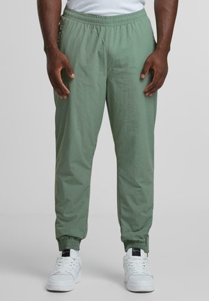 Donkerharige persoon die lichtgroene broek met elastische taille en witte sneakers draagt, staand tegen een grijze achtergrond.