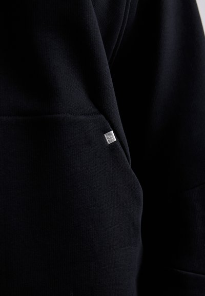 Črna majica iz puloverja z teksturirano površino, ki ima majhno blagovno znamko v srebrni barvi na žepu. Preprost dizajn brez vzorcev.