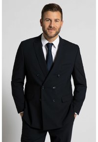 Zwarte dubbelbreasted suit met gekartelde revers, met twee voorzakken en blauwe knopen. Een wit shirt en een donkere stropdas maken de look compleet.