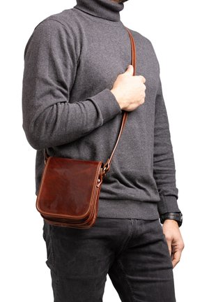 Man draagt een grijze coltrui en zwarte broek, houdt een bruine leren crossbodytas vast aan het hengsel op een witte achtergrond.