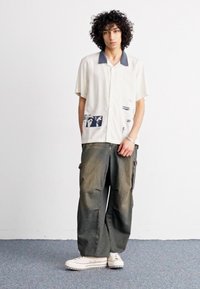 Camicia crema a maniche corte con colletto blu e stampe astratte, abbinata a pantaloni verde scuro larghi e leggermente usurati e sneakers bianche.
