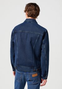 Homme aux cheveux ondulés portant une veste en denim bleu foncé et un jean bleu avec un patch en cuir Wrangler, vu de dos sur un fond uni.
