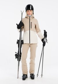 Ski apranga, apimanti kreminės ir baltos spalvos striukę, kremine kelnes, juodus pirštines ir šalmą. Laiko slidės ir slidžių lazdas. Matomi tekstūruoti audiniai.