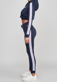 Žena v námořnické modré zkrácené mikině a odpovídajících teplákách s bílými postranními pruhy a teniskách, stojící bokem k šedému pozadí.