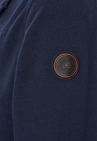 Marineblauer, gerippter Sweatshirt mit einem runden schwarzen Patch mit roten Akzenten und Logo auf dem Ärmel. Hergestellt aus strukturiertem Stoff für maximalen Komfort.