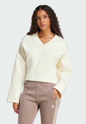 Cream V-neck sweatshirt with a cropped design and wide sleeves; features subtle embossed logo. Paired with light brown tapered pants with stripes.
