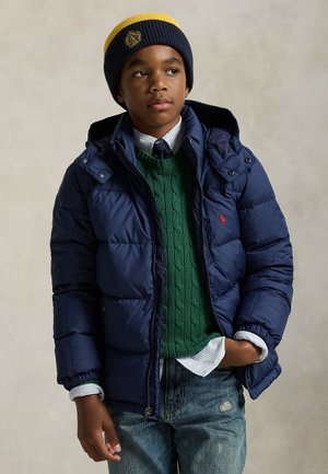 Junge trägt navyblaue Steppjacke, grünen Zopfpullover, gestreiftes Hemd, Krawatte, blaue und gelbe Mütze und abgenutzte Jeans mit einer Hand in der Tasche.